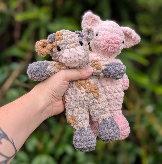 A crocheted mini Cow and pig snuggler held up against a leafy background