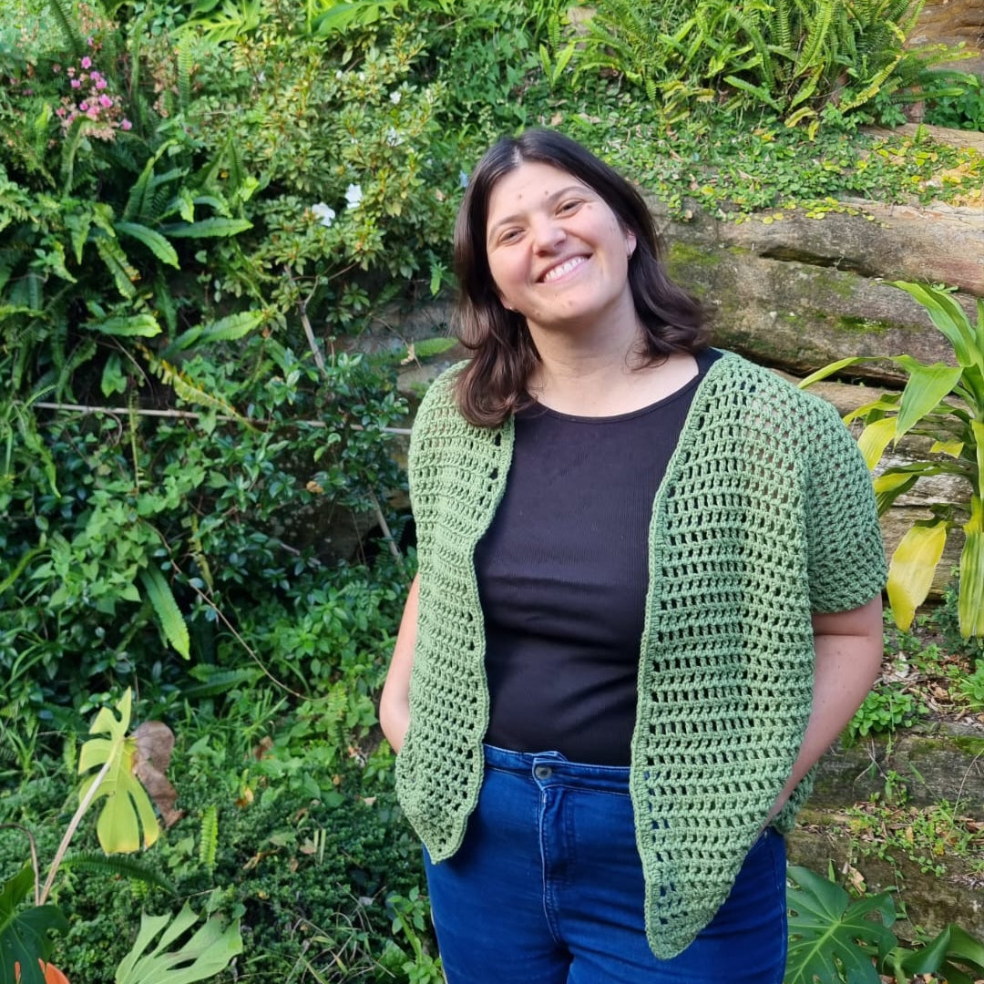 A person in blue jeans, a balck singlet, and a green handmade crochet mesh shirt stands in front of greenery.