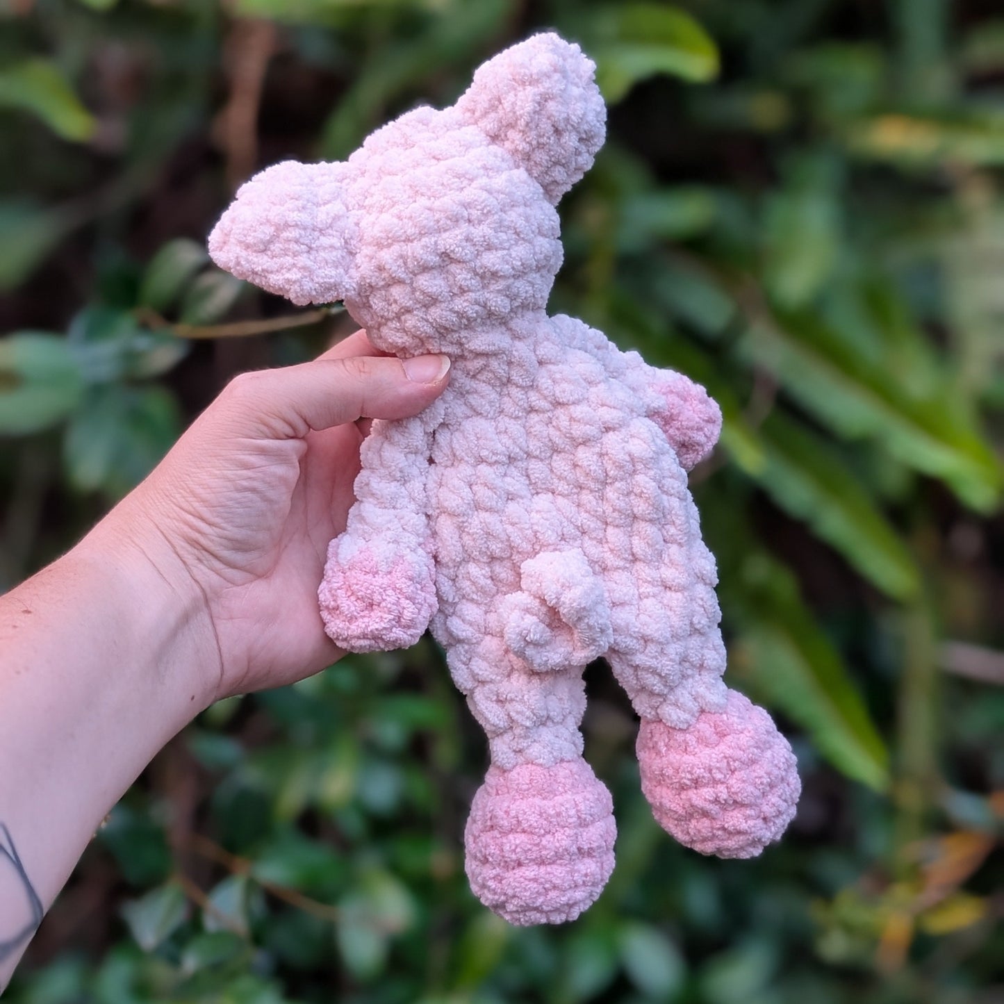 Hand holding a pink crochet toy against a green leafy background