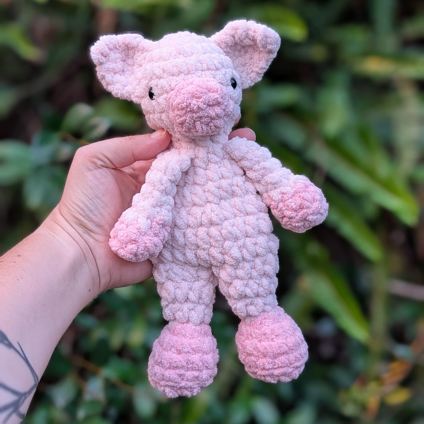 Hand holding a pink crochet pig toy against a green leafy background