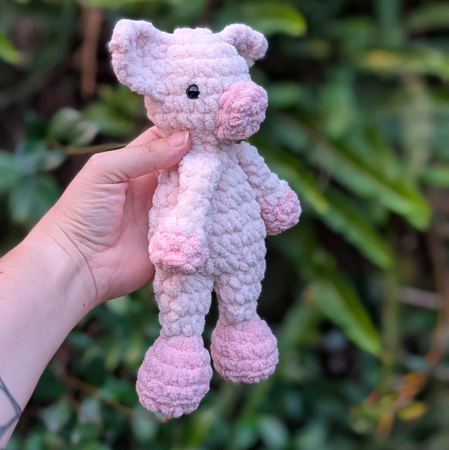 Hand holding a small pink crochet pig against a green leafy background