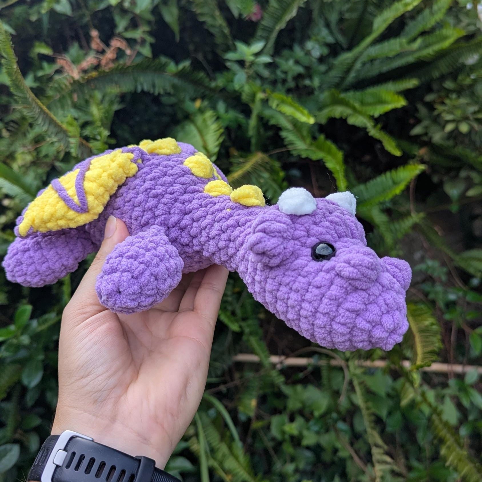 Crocheted dragon facing the camera - it's body is purple and it has white horns with yellow spikes and wings.